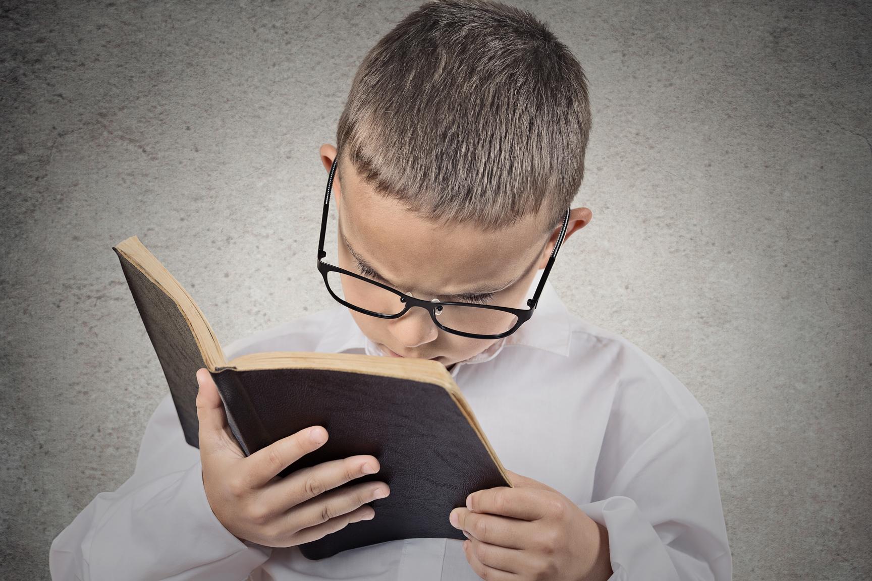 Close up portrait young, little man with glasses holding, reading old book, having difficulty to see text isolated grey wall background. Face expression. Education concept. Vision problems in children