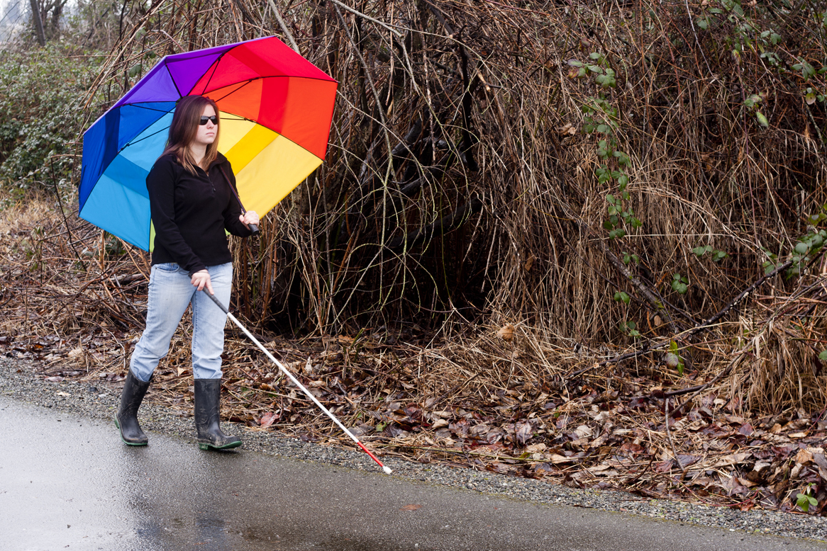 A cane user feels her way along a park trail while carrying a brightly colored umbrella in the rain.