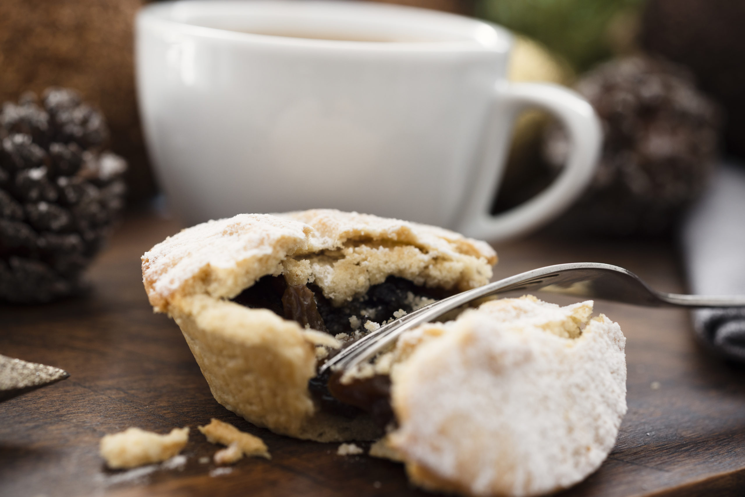 Close up of a mince pie split open with fork dusted with icing sugar, served with a cup of coffee with christmas decorations in the background. Festive Christmas food scene.