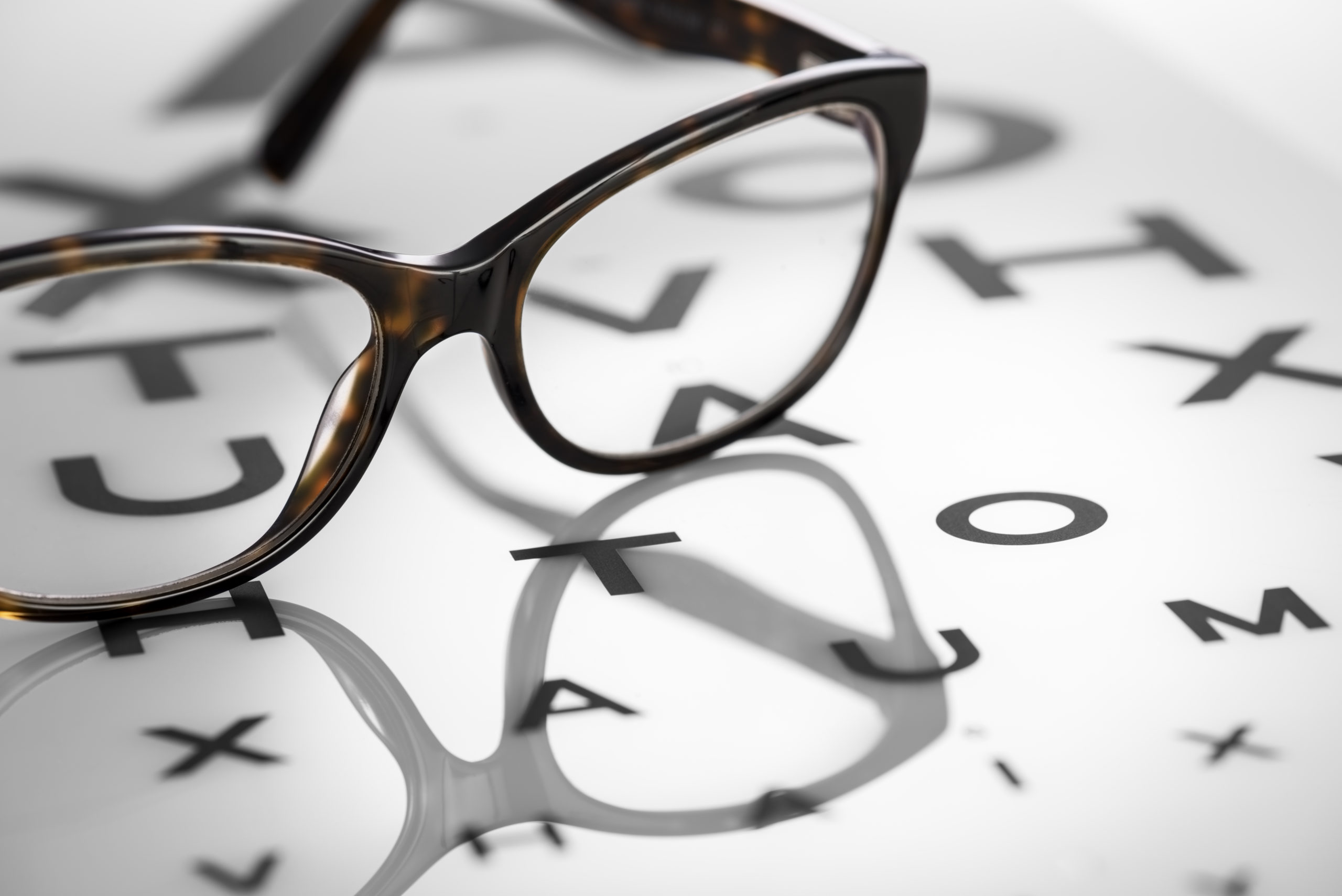 Brown plastic reading spectacles are lying on an alphabetic sight chart. The chart is made of thick white plastic with black letters that get smaller in size as they go down the chart. There is a pool of light surrounding the glasses.